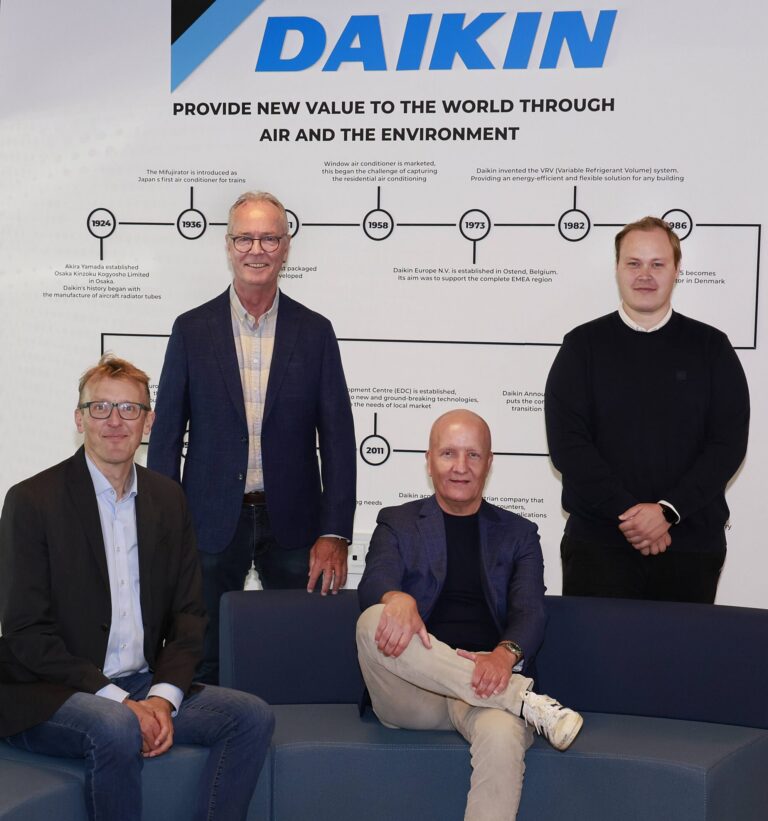 Fire mænd — to siddende på en blå sofa og to stående bag dem — poserer foran en væg med firmalogoet "DAIKIN" og slagordet "PROVIDE NEW VALUE TO THE WORLD THROUGH AIR AND THE ENVIRONMENT".