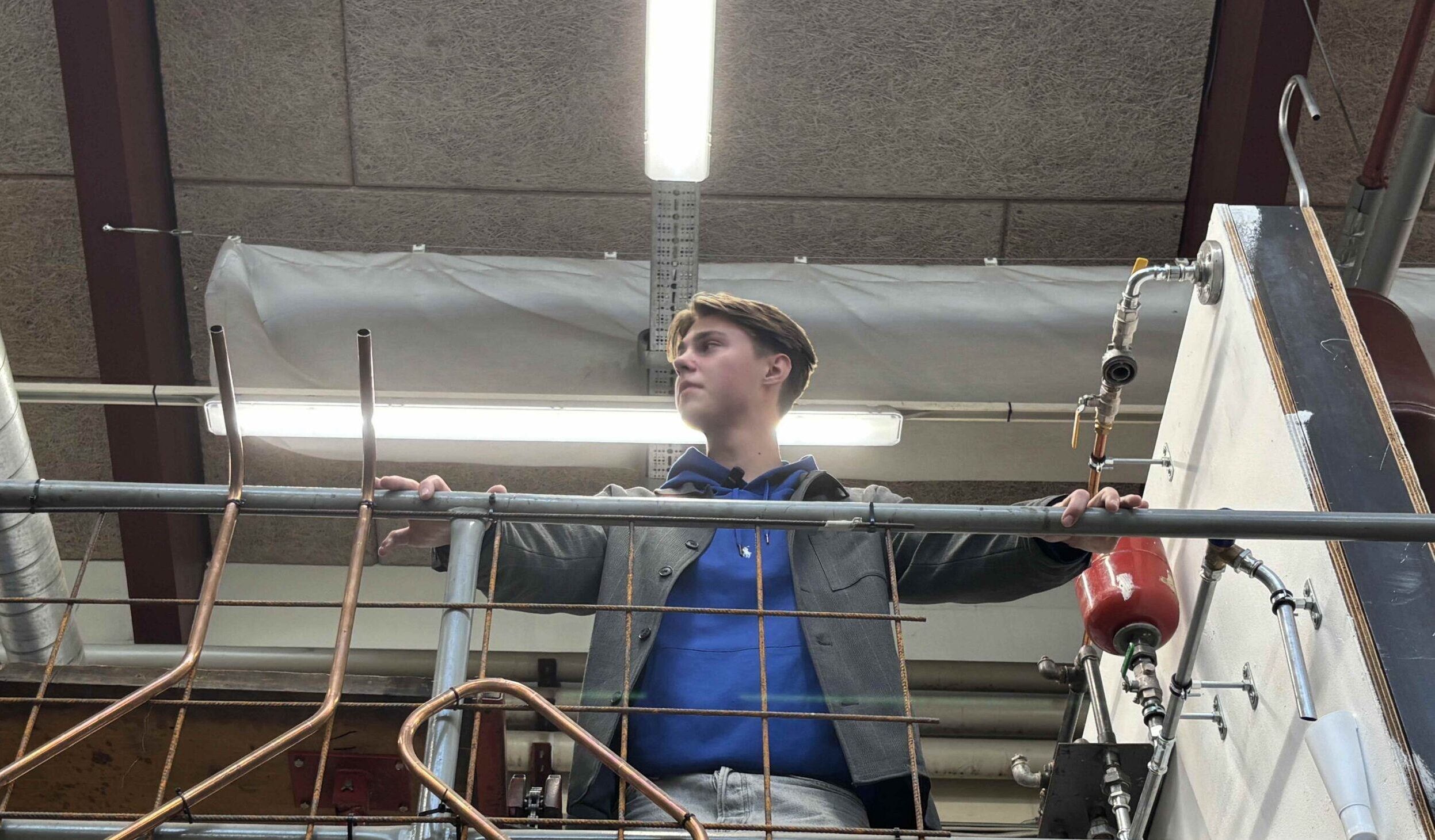 Ung person iført blå hættetrøje og grå jakke står med armene udstrakte og holder ved et metalgelænder på en forhøjning i et industriværksted omgivet af kobberrør, ventiler og loftsbelysning.