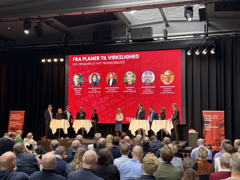 Paneldebat på en scene med ni paneldeltagere stående ved høje runde borde foran et stort rødt skærm og et fyldt publikum i forgrunden. På skærmen står teksten "FRA PLANER TIL VIRKELIGHED" og "NYE OPGAVER I ET NYT TRUSSELSBILLEDE", der vises også cirkulære portrætter med navne; på venstre banner står "Faglærte får installationerne til at fungere. Også når vi har mest brug for det." og på højre banner står "Strøm, vand og varme er kritisk. Det er fagligheden også."