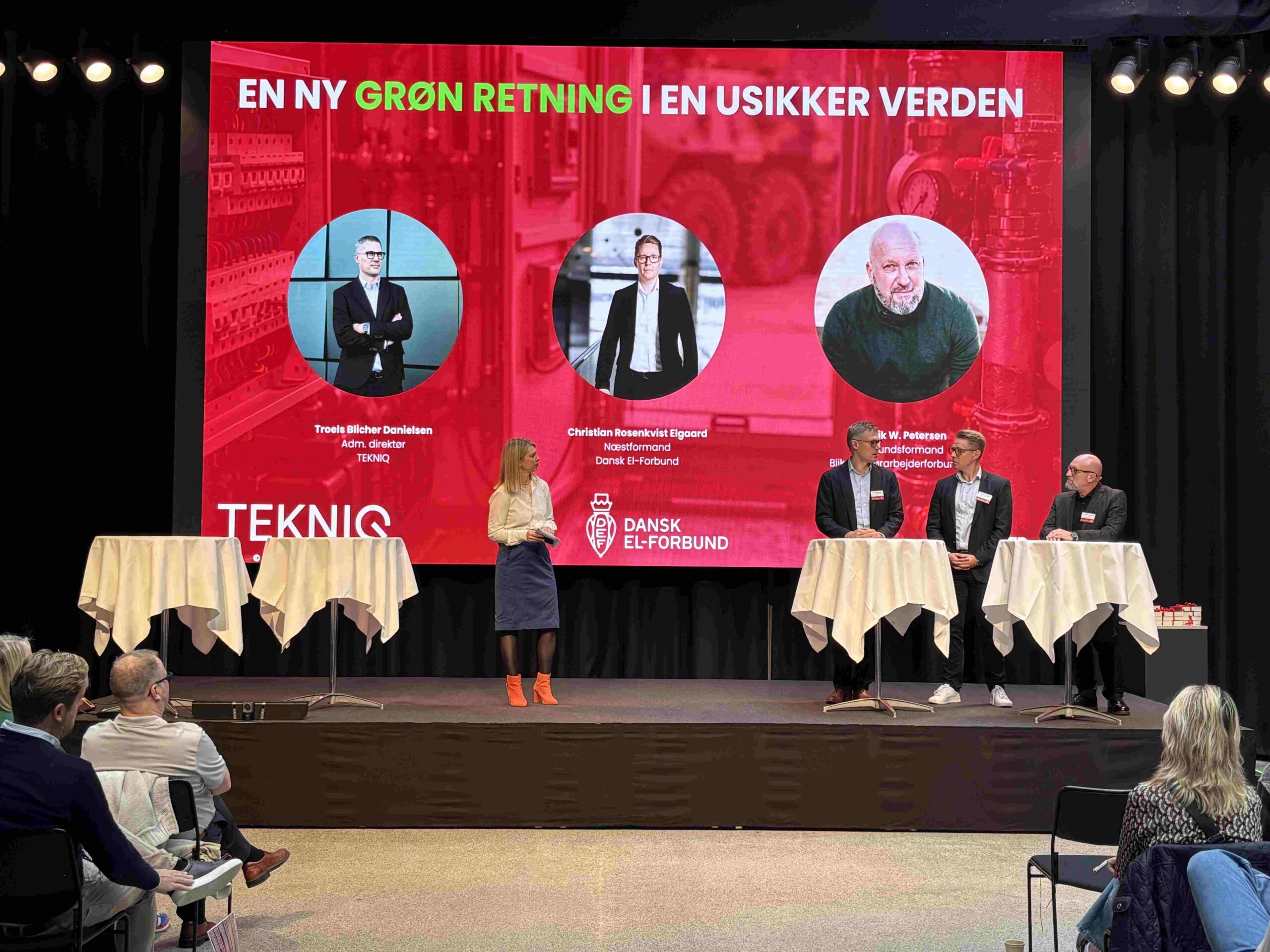 Fire talere står på en scene foran en stor rød skærm med teksten "EN NY GRØN RETNING I EN USIKKER VERDEN"; skærmen viser tre portrætbilleder med navne og titler under hvert samt logoerne "TEKNIQ" og "DANSK EL-FORBUND".