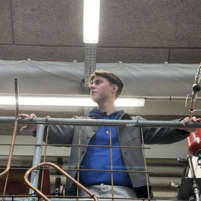 Ung person iført blå hættetrøje og grå jakke står med armene udstrakte og holder ved et metalgelænder på en forhøjning i et industriværksted omgivet af kobberrør, ventiler og loftsbelysning.